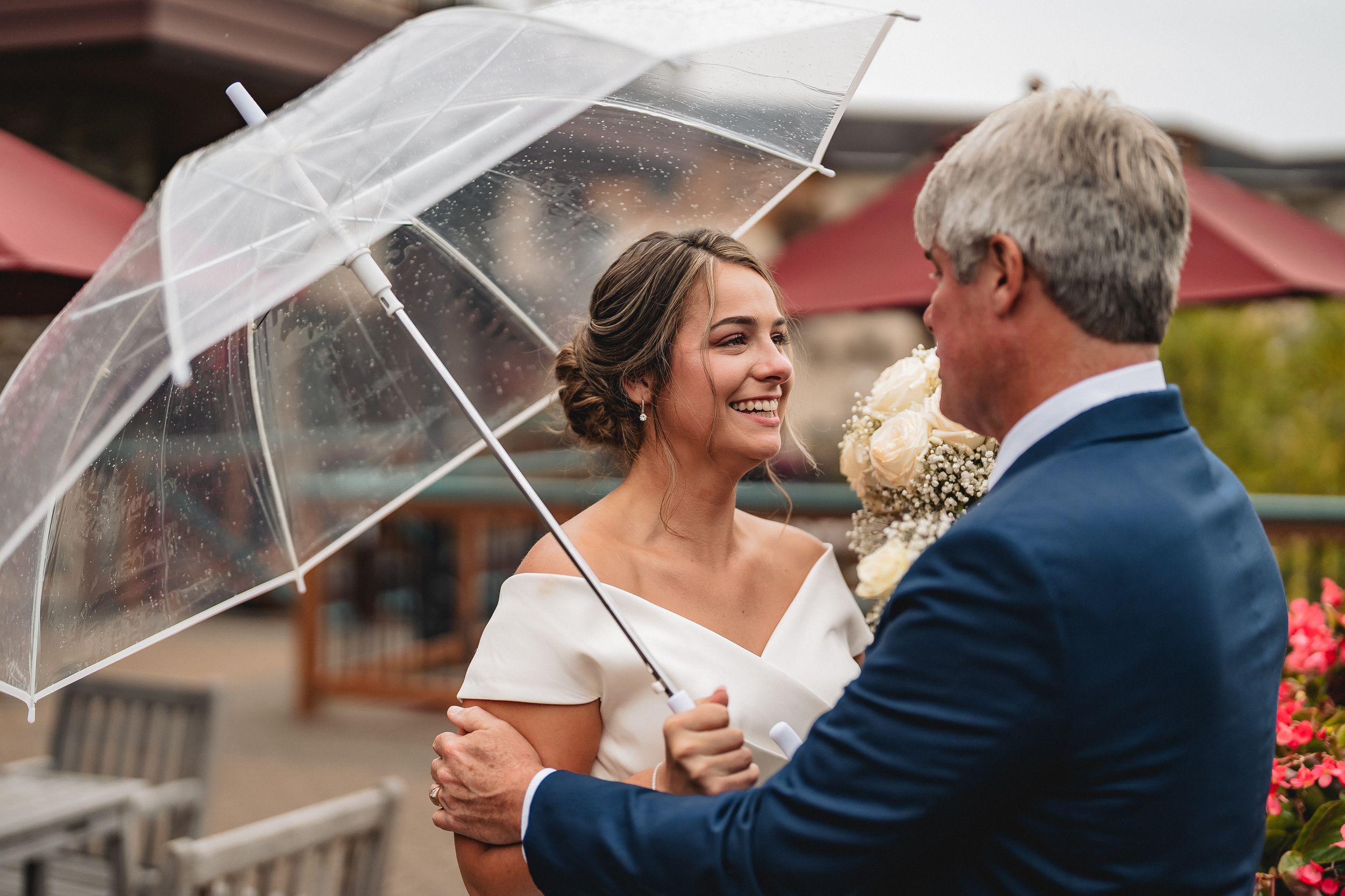 Two people share an intimate moment under a clear umbrella surrounded by a garden setting.