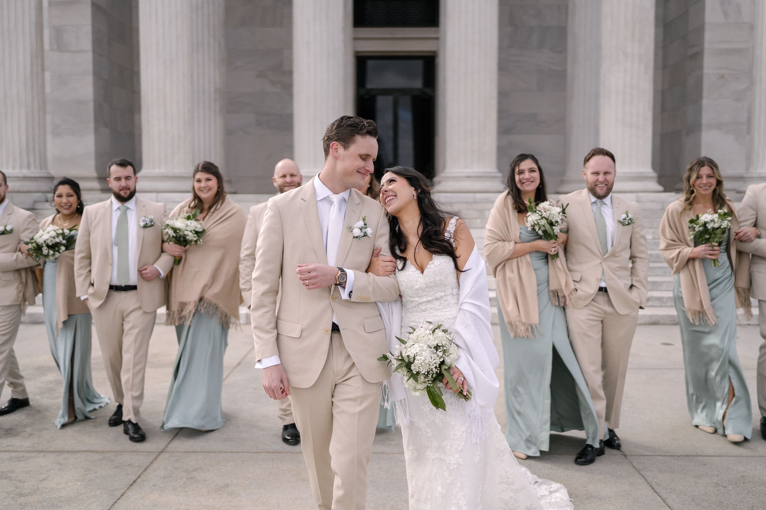 Wedding party in light beige and blue attire poses on elegant stone steps with classic columns in background.