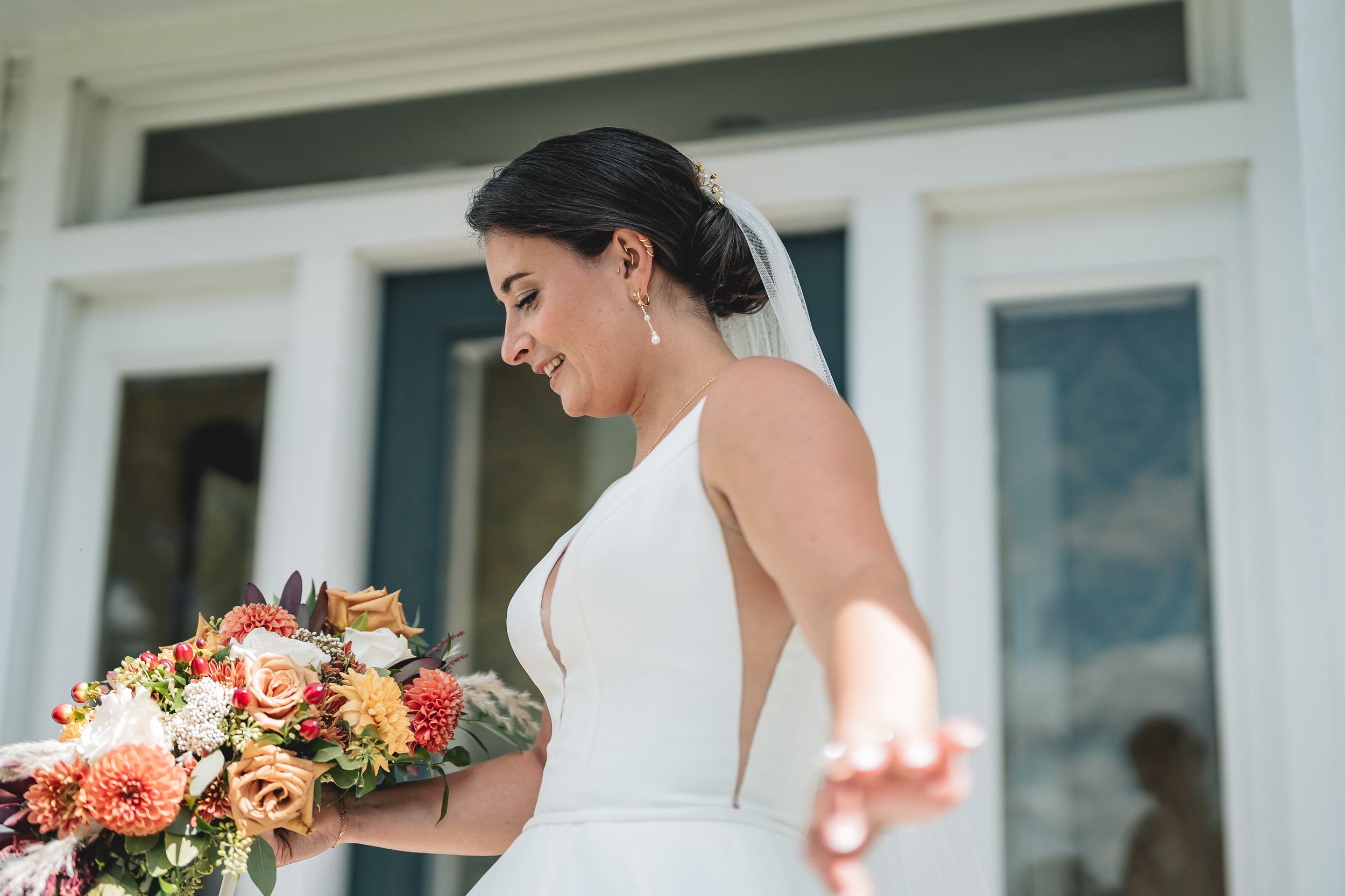 Beautiful bride in elegant halter wedding gown holds peach and orange floral bouquet while smiling outdoors.