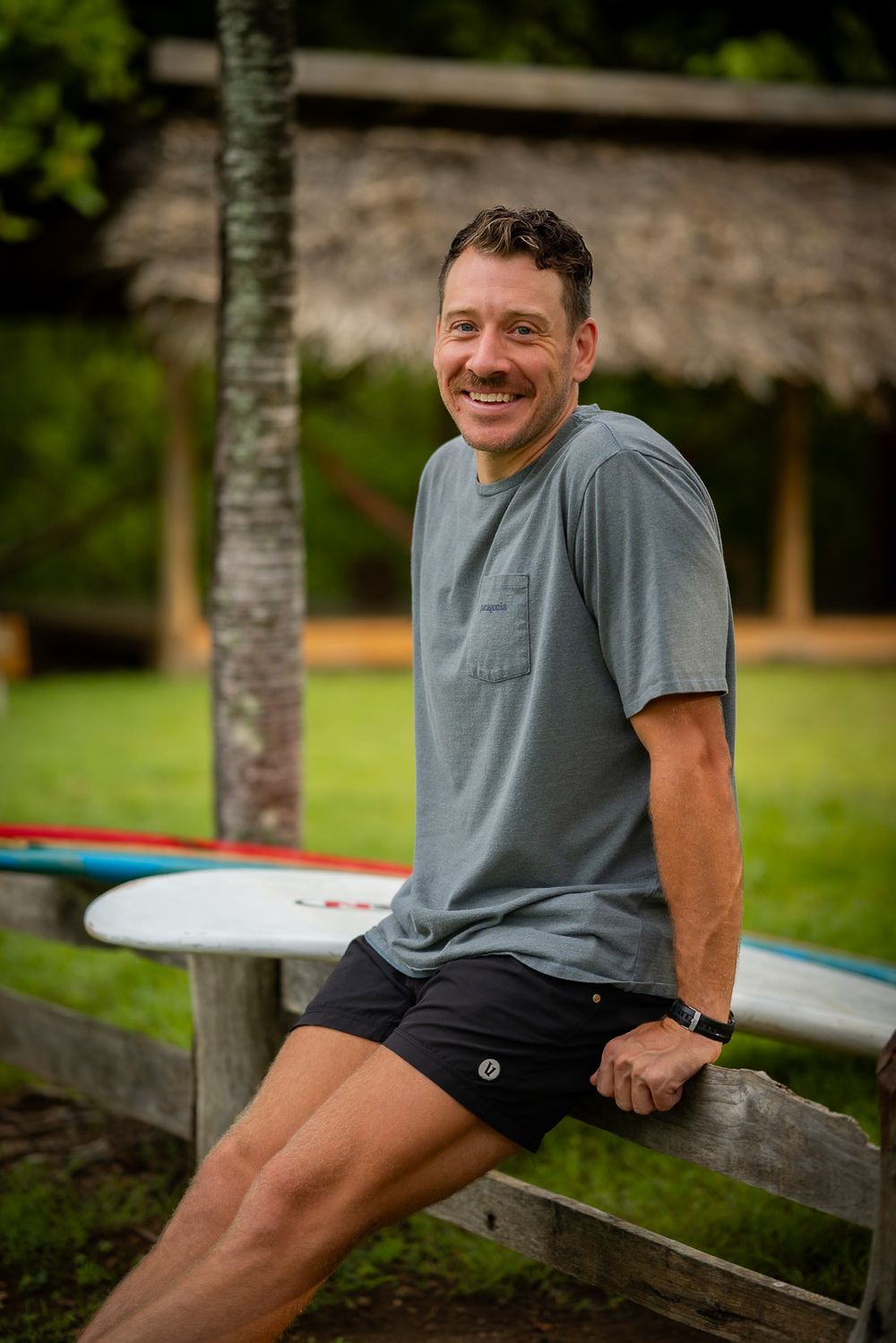 Person sitting on a wooden fence with a surfboard in the background.
