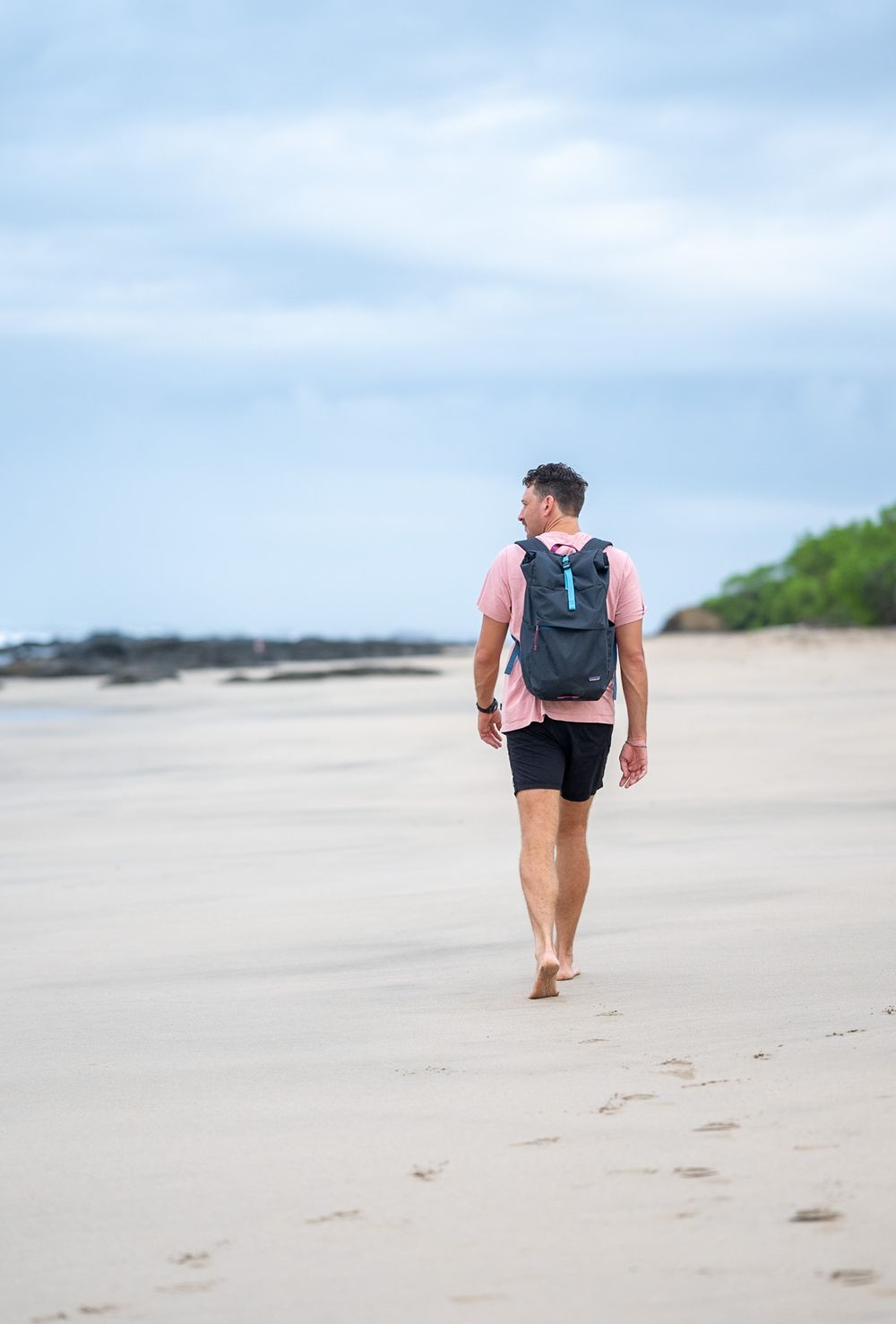 Figure walking alone on a sandy beach with a backpack against cloudy sky.