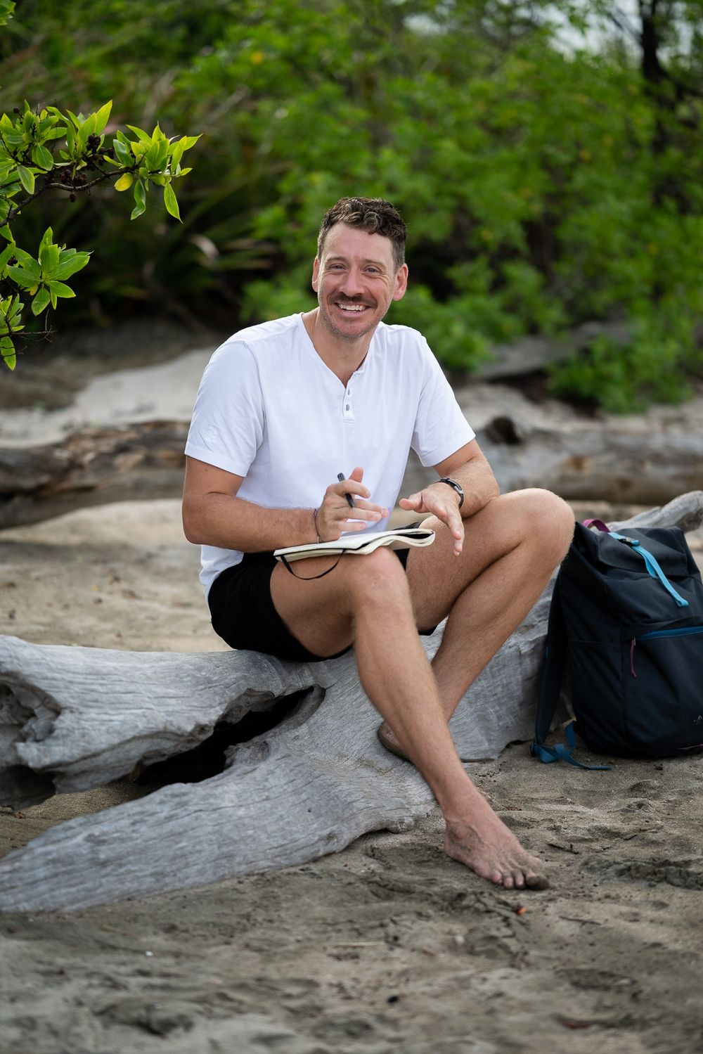 Person writing in notebook while sitting on beach rocks surrounded by greenery.