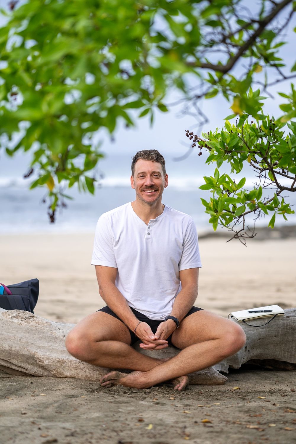 Person in white shirt sits cross-legged and smiling on beach with lush green foliage overhead.