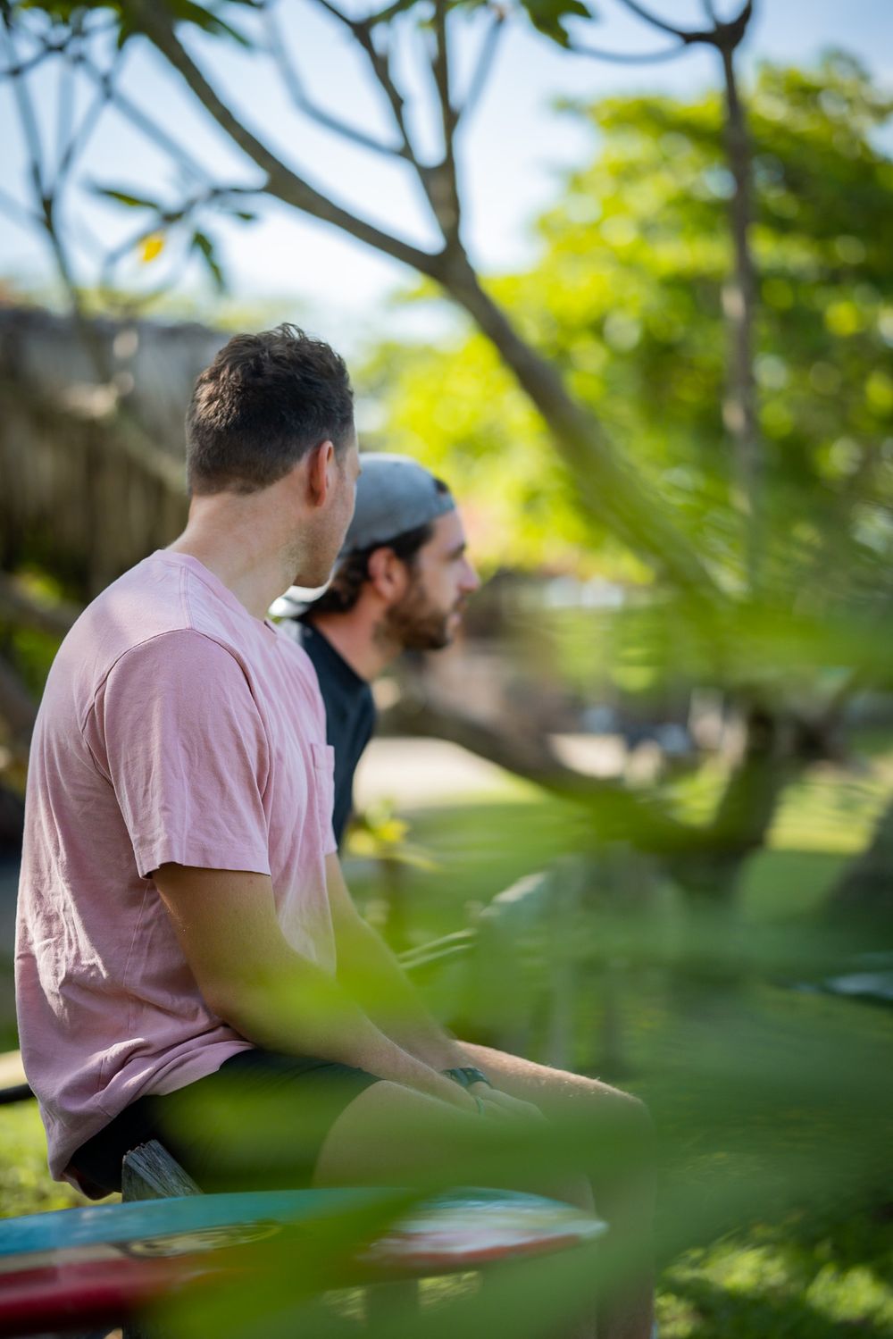 Two people sitting outdoors amid tropical foliage with surfboards nearby.