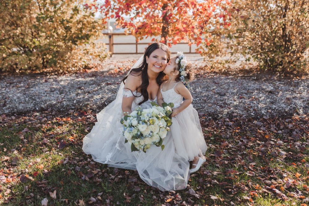 A bride in a white dress holds a bouquet while kneeling on autumn leaves with two small dogs.