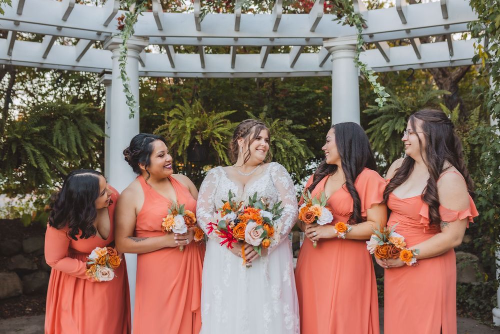 Bride in white lace gown stands with bridesmaids wearing coral dresses holding orange and white floral bouquets under pergola.