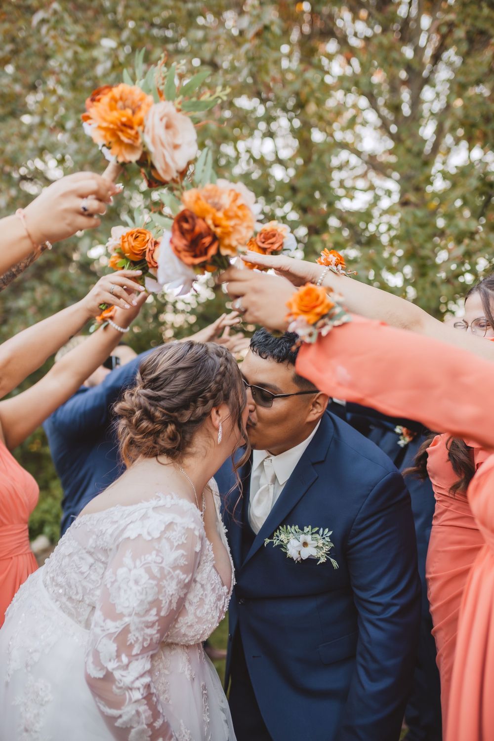 Wedding celebration with bridesmaids holding coral and peach roses overhead as couple shares intimate moment outdoors.