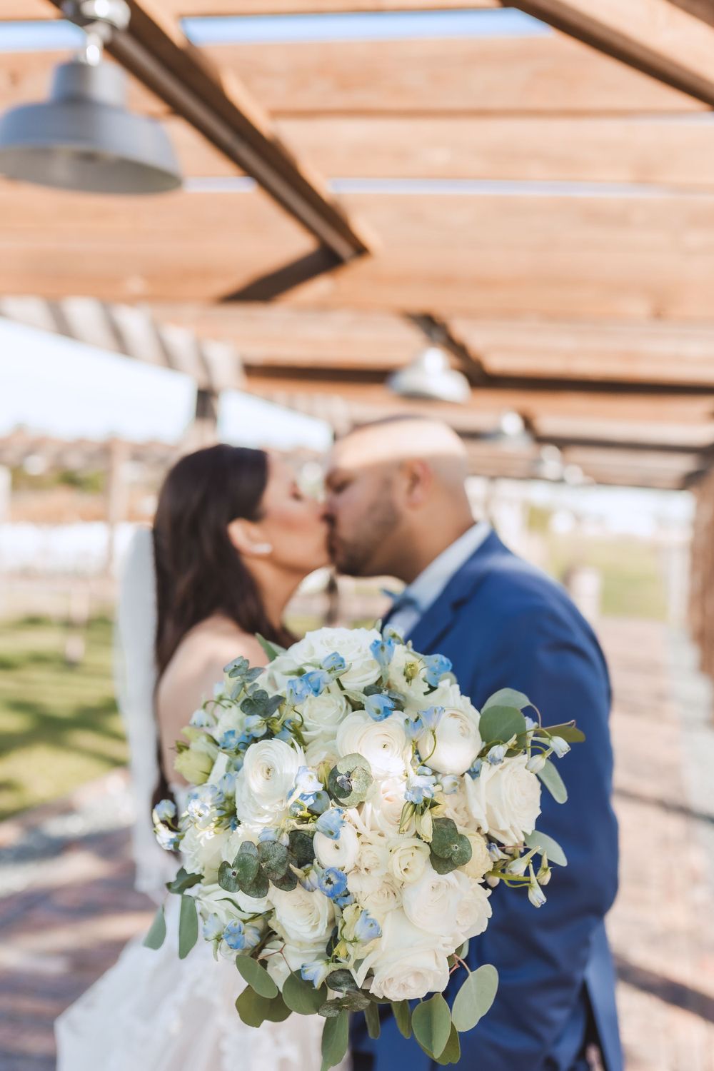 A lovely white and eucalyptus wedding bouquet in focus while a couple shares an intimate moment under a wooden pergola.