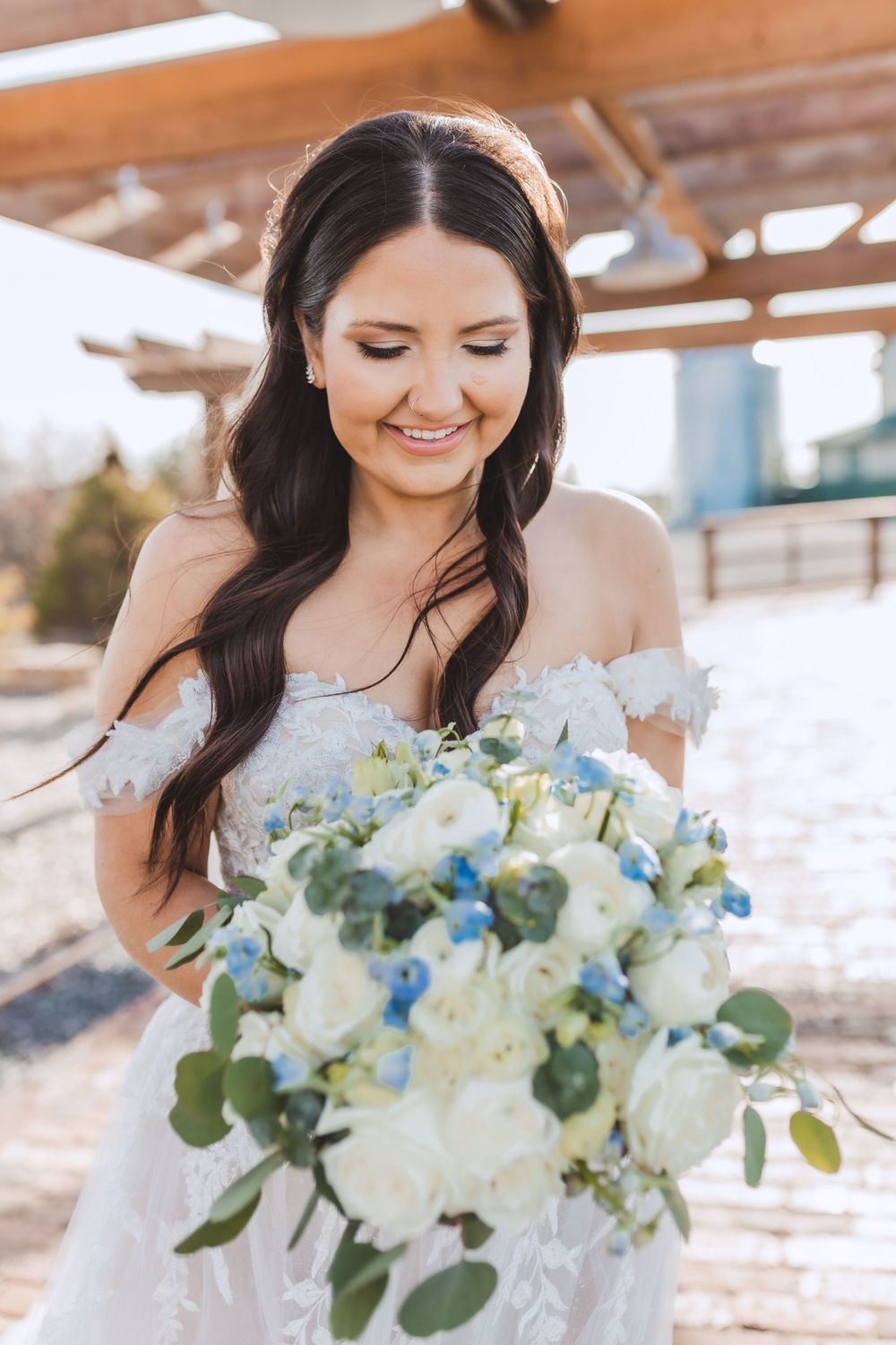 A bride in an off-shoulder lace dress smiles while holding a bouquet of white roses and blue flowers with eucalyptus.