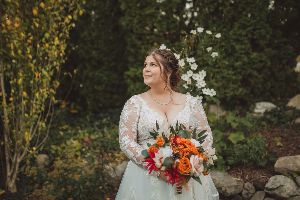 A bride in a white lace gown holds a vibrant orange and pink bouquet in a garden setting.