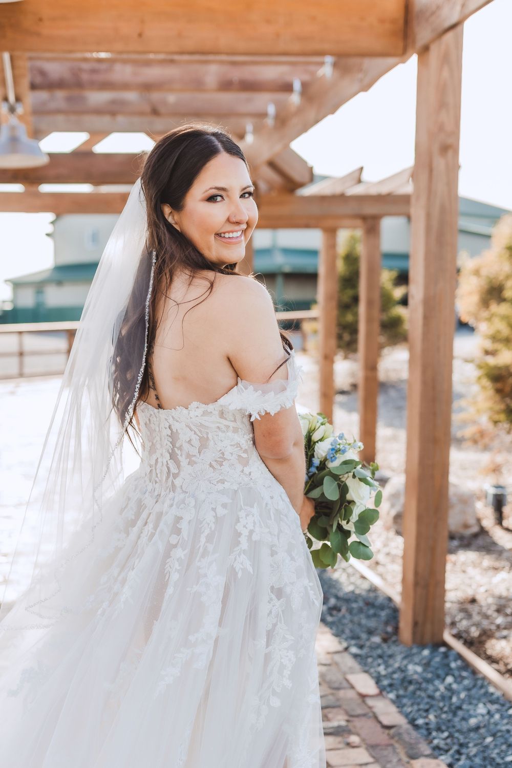 A bride in a lace off-shoulder wedding gown smiles while holding a bouquet under a wooden pergola.