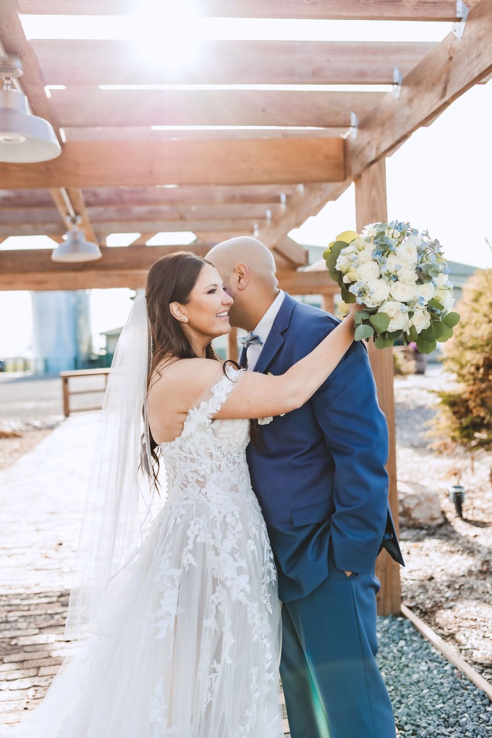 A couple embraces under a wooden pergola, with the bride in a lace gown and groom in navy suit holding a white bouquet.