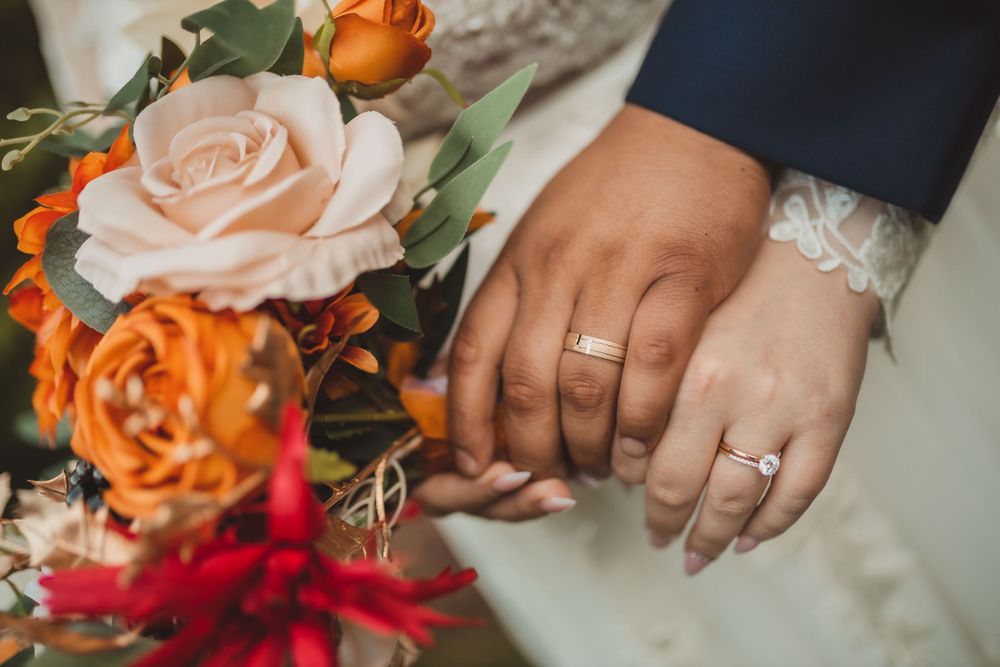 Close-up of wedding rings and hands beside a bouquet featuring peach and orange roses with red floral accents.