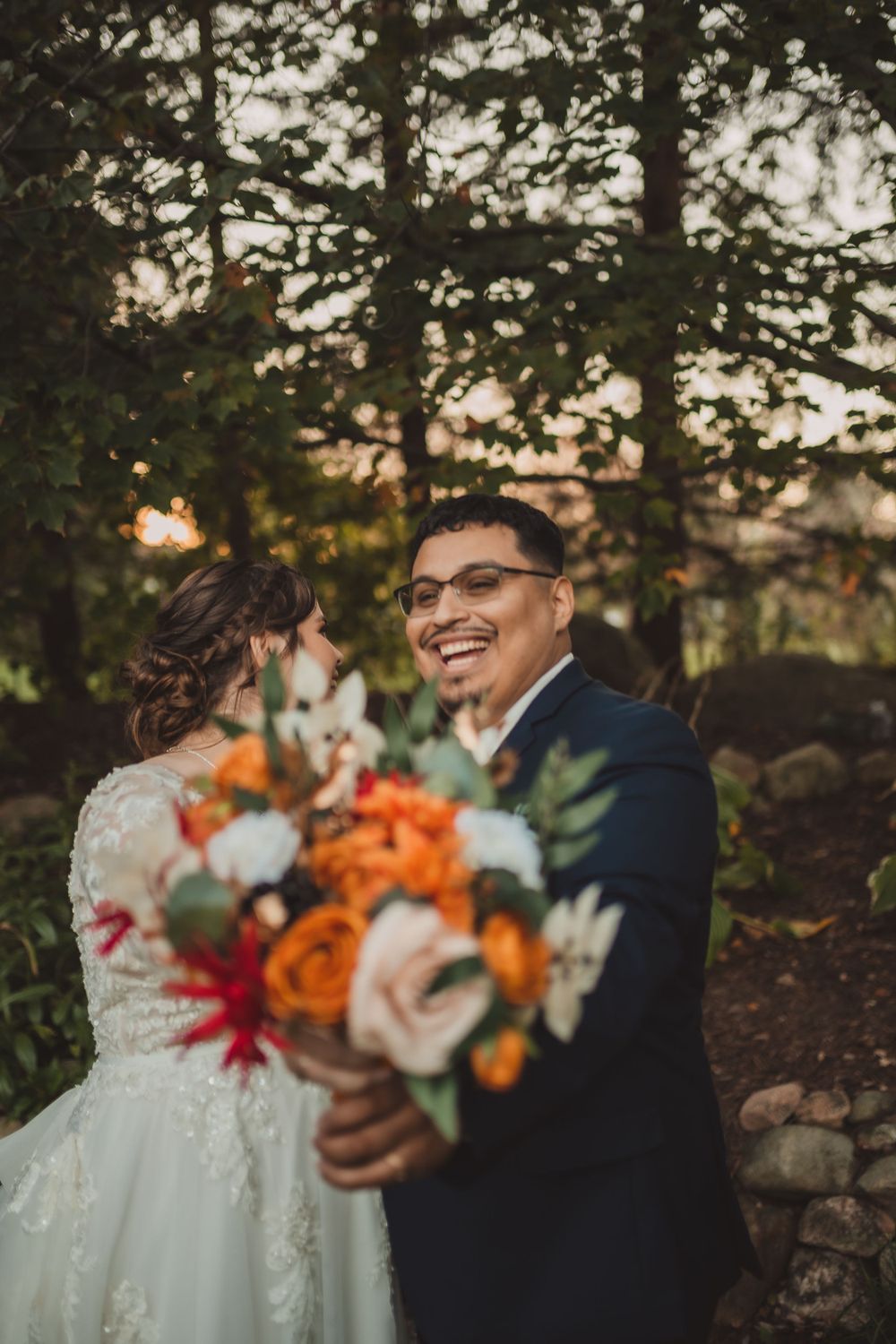 A joyful outdoor wedding portrait features a beautiful bouquet with orange and white flowers against a natural sunset backdrop.
