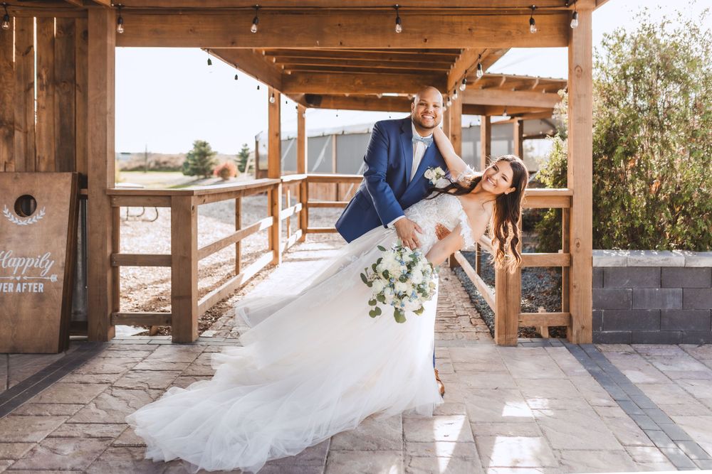 Couple shares a romantic dip pose under rustic wooden pergola at outdoor wedding venue in natural setting.