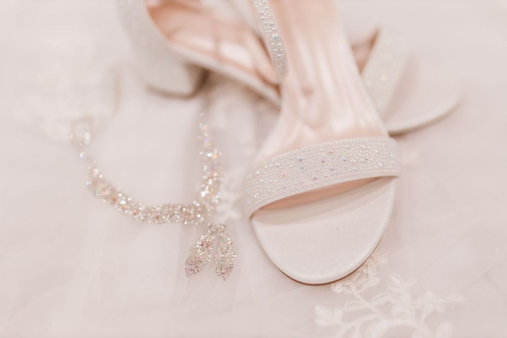 A delicate crystal necklace rests beside blush pink bridal heels on soft white fabric.