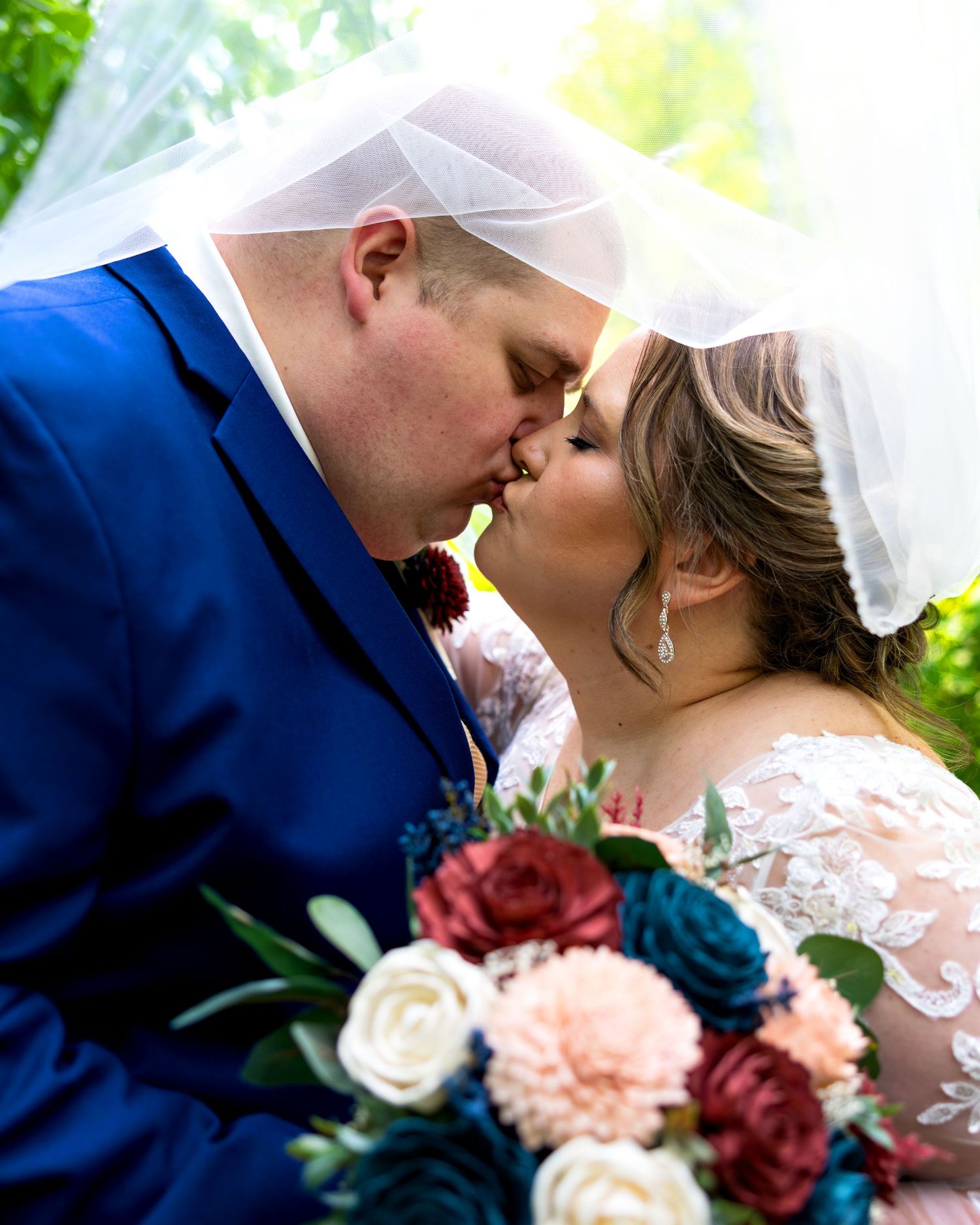 Bride and groom sharing romantic kiss under wedding veil with colorful bridal bouquet in foreground.