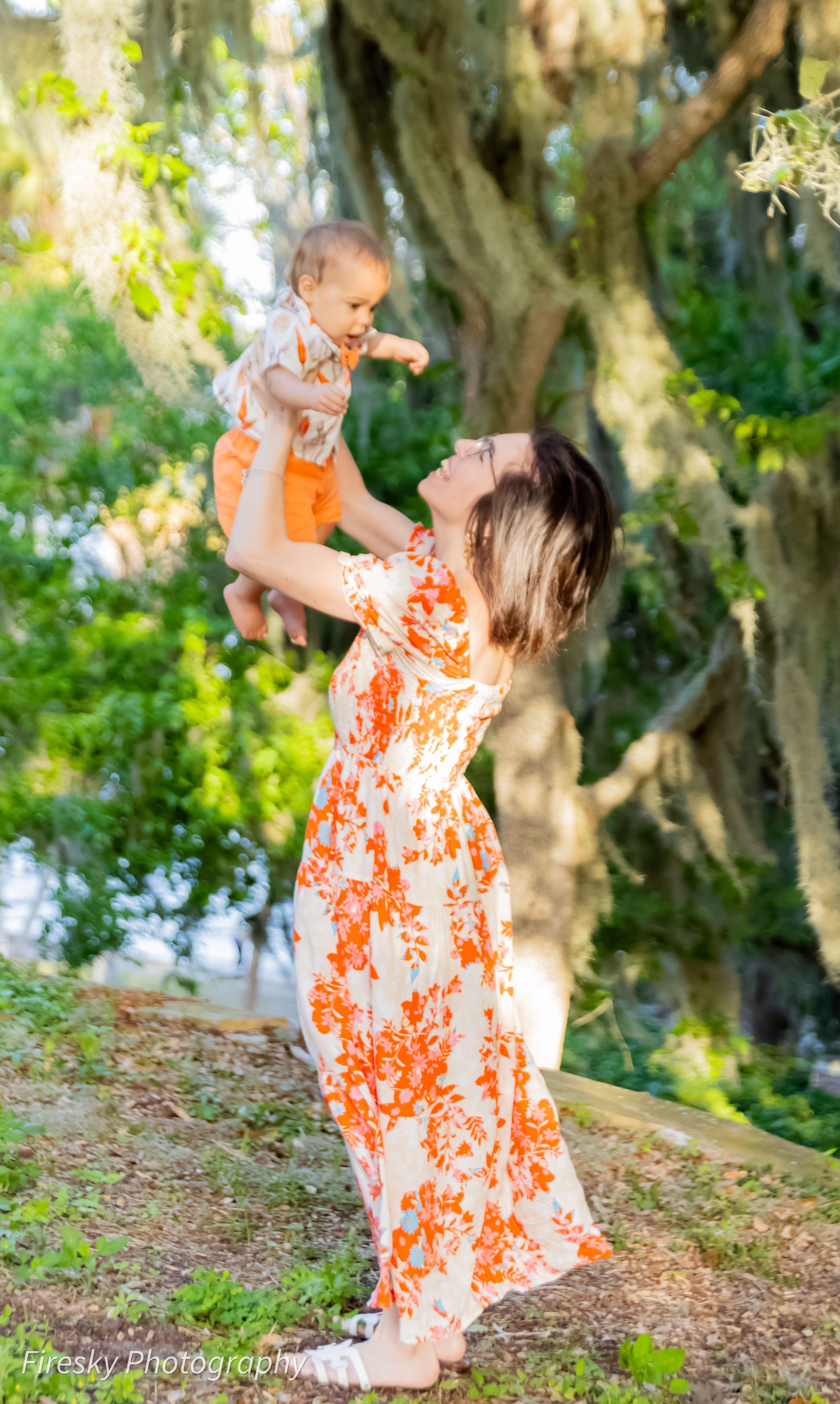 A woman in an orange floral dress lifts a baby high in the air against a garden backdrop.