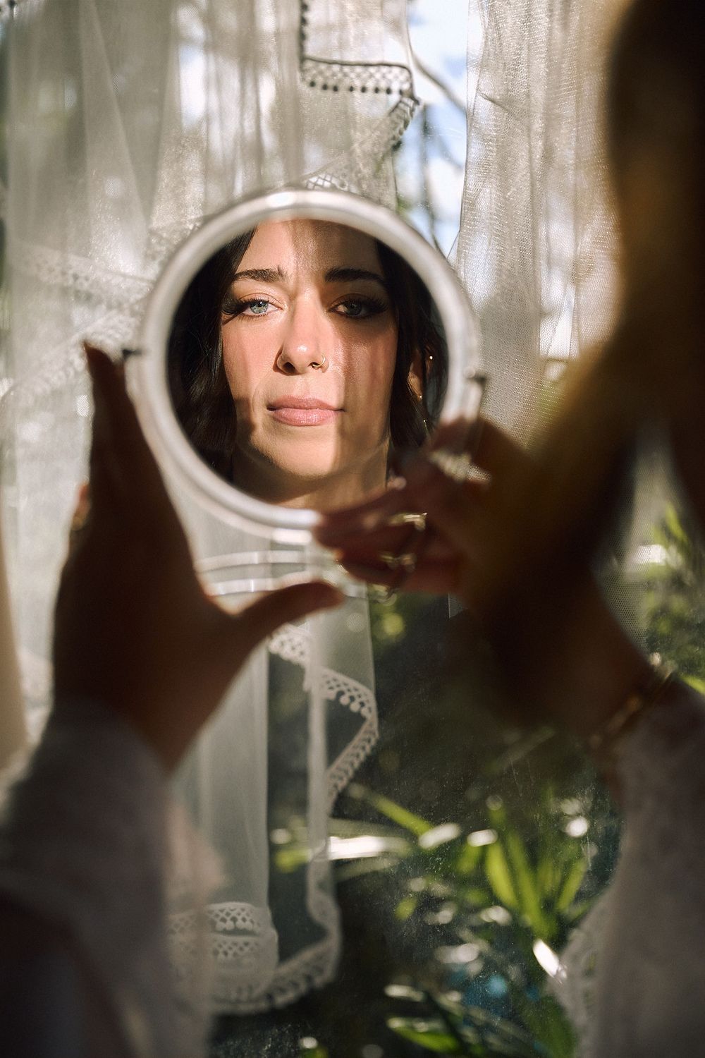 A woman gazes at her reflection in a round handheld mirror, framed by sheer white curtains in soft sunlight.