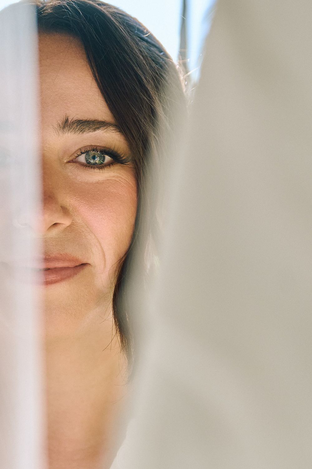 Close-up portrait of a smiling brunette woman with green eyes, partially hidden behind a white sheer curtain.