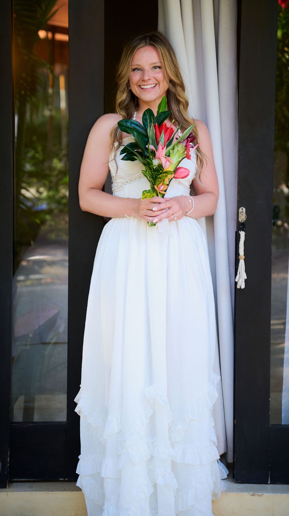 Smiling bride in white ruffled gown holding tropical floral bouquet standing in doorway.