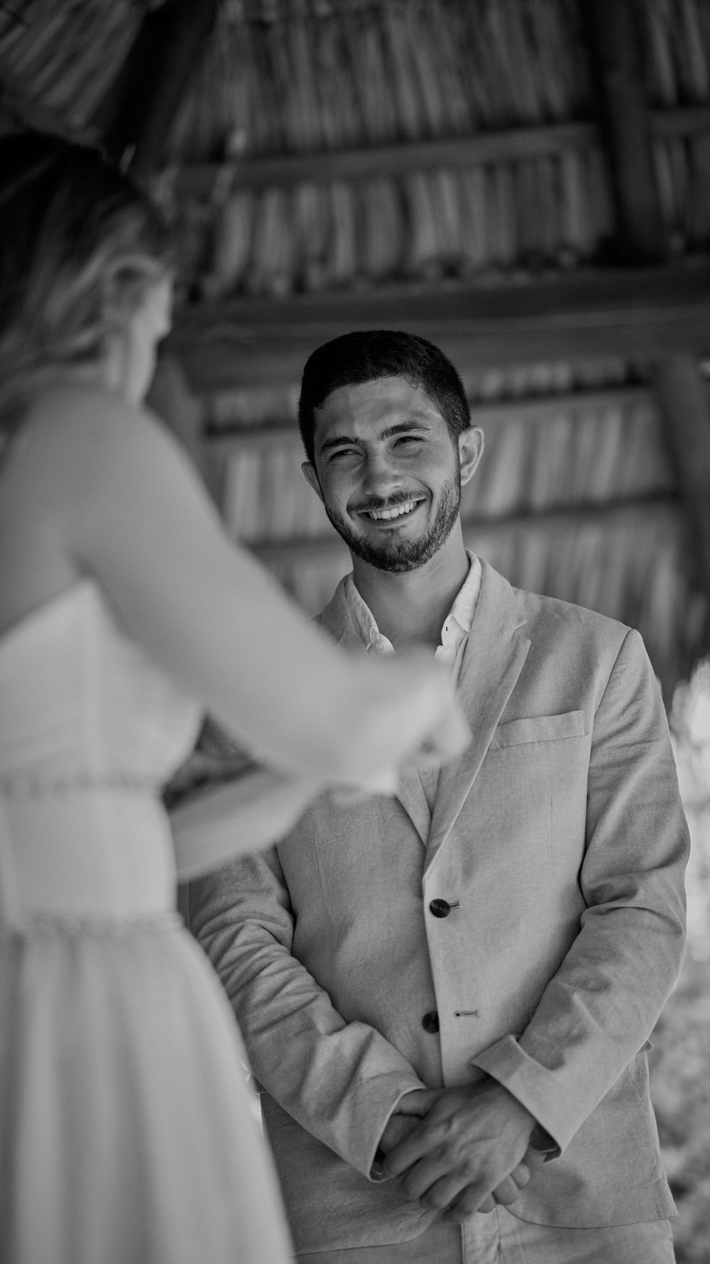 Groom smiling joyfully at bride during outdoor wedding ceremony under thatched roof in black and white photo.