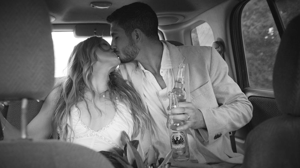 Black and white photo of a couple in wedding attire sharing a romantic kiss in a car, holding champagne.