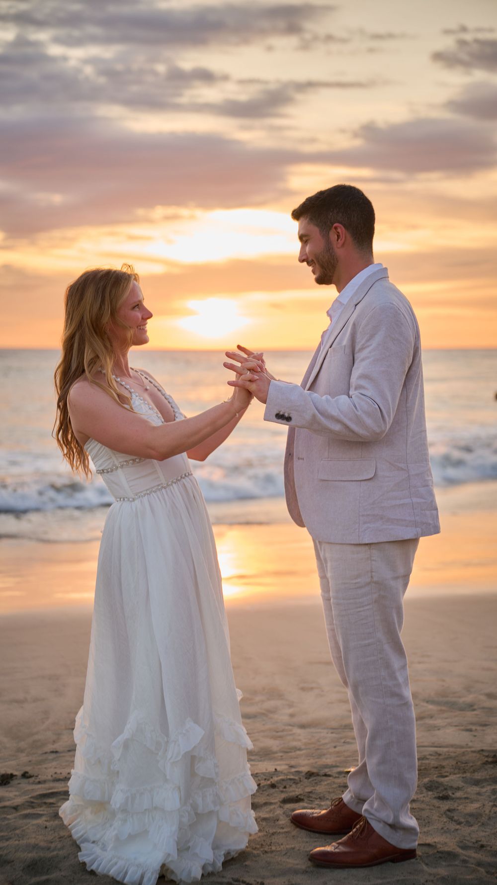 Couple exchanging rings on a beach at sunset, bride in white gown and groom in light suit, ocean waves behind them.