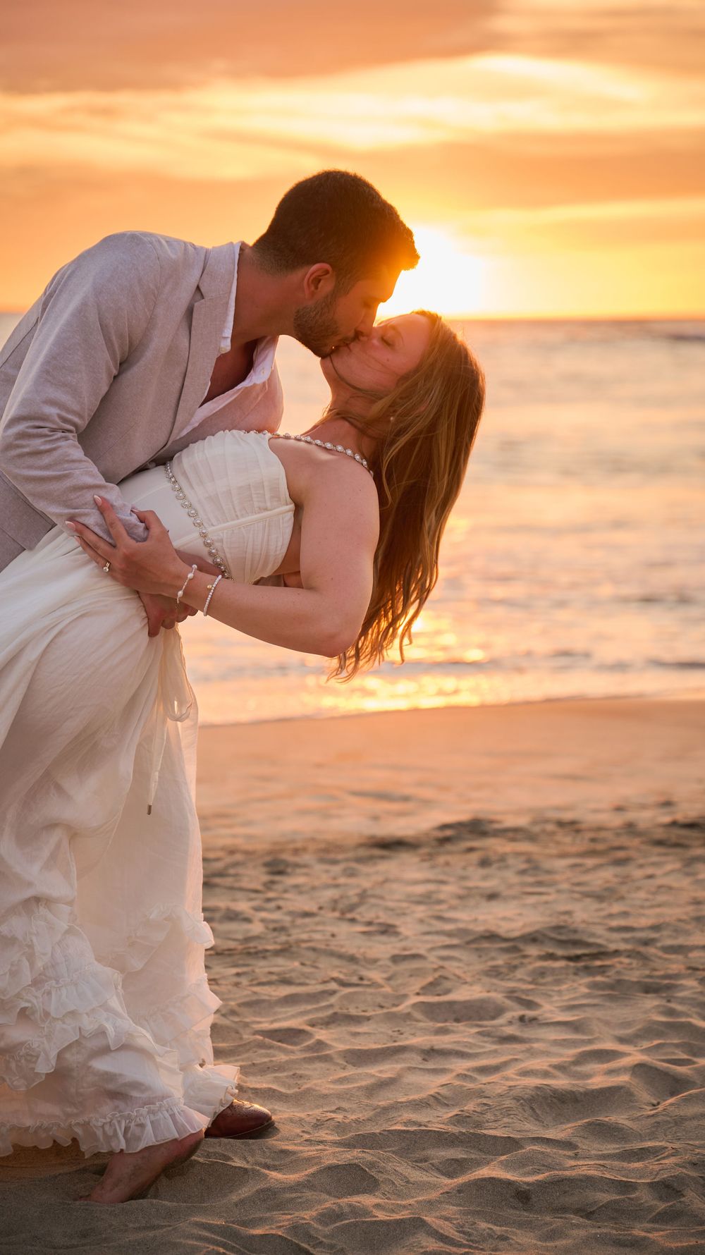A couple shares a romantic dip kiss on a sandy beach at golden sunset, bride in white dress.