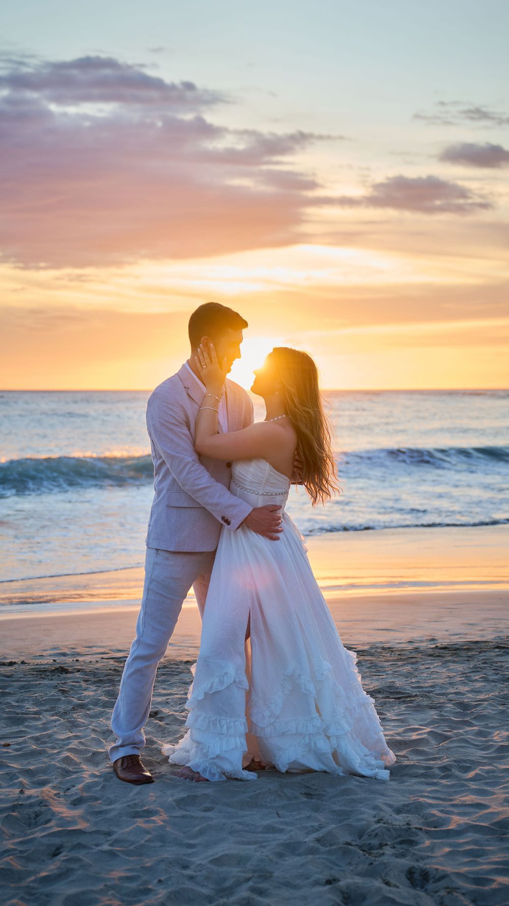 Couple in wedding attire embracing on a sandy beach at golden sunset with ocean waves in the background.