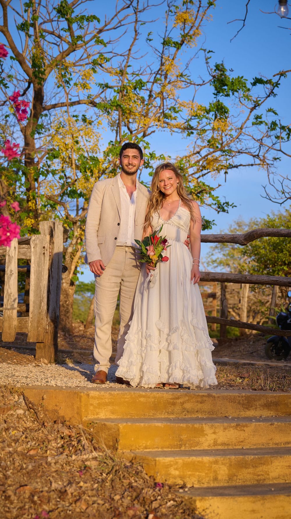 Couple in wedding attire standing on stone steps outdoors, bride holding tropical bouquet, groom in beige suit, at sunset.