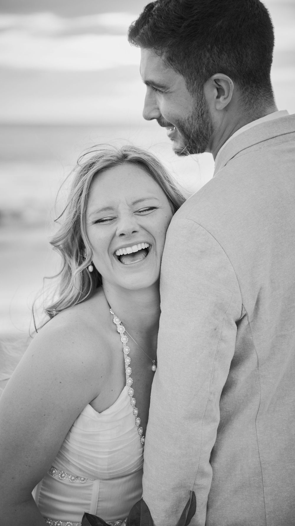 Black and white photo of a laughing couple embracing on a beach, woman in white dress smiling joyfully at man in suit.