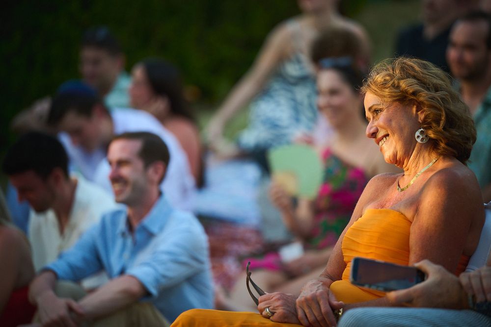 A smiling woman in an orange dress sits outdoors at a social gathering with laughing guests in the background.