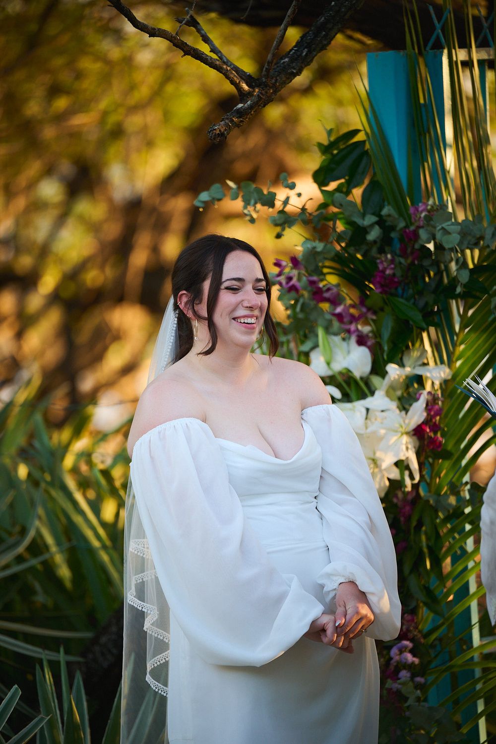 Smiling bride in off-shoulder white gown with veil stands at outdoor ceremony altar surrounded by lush greenery.