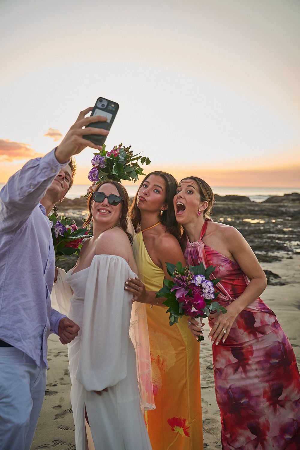 Group of women in colorful dresses taking a selfie on a beach at sunset during a wedding celebration.