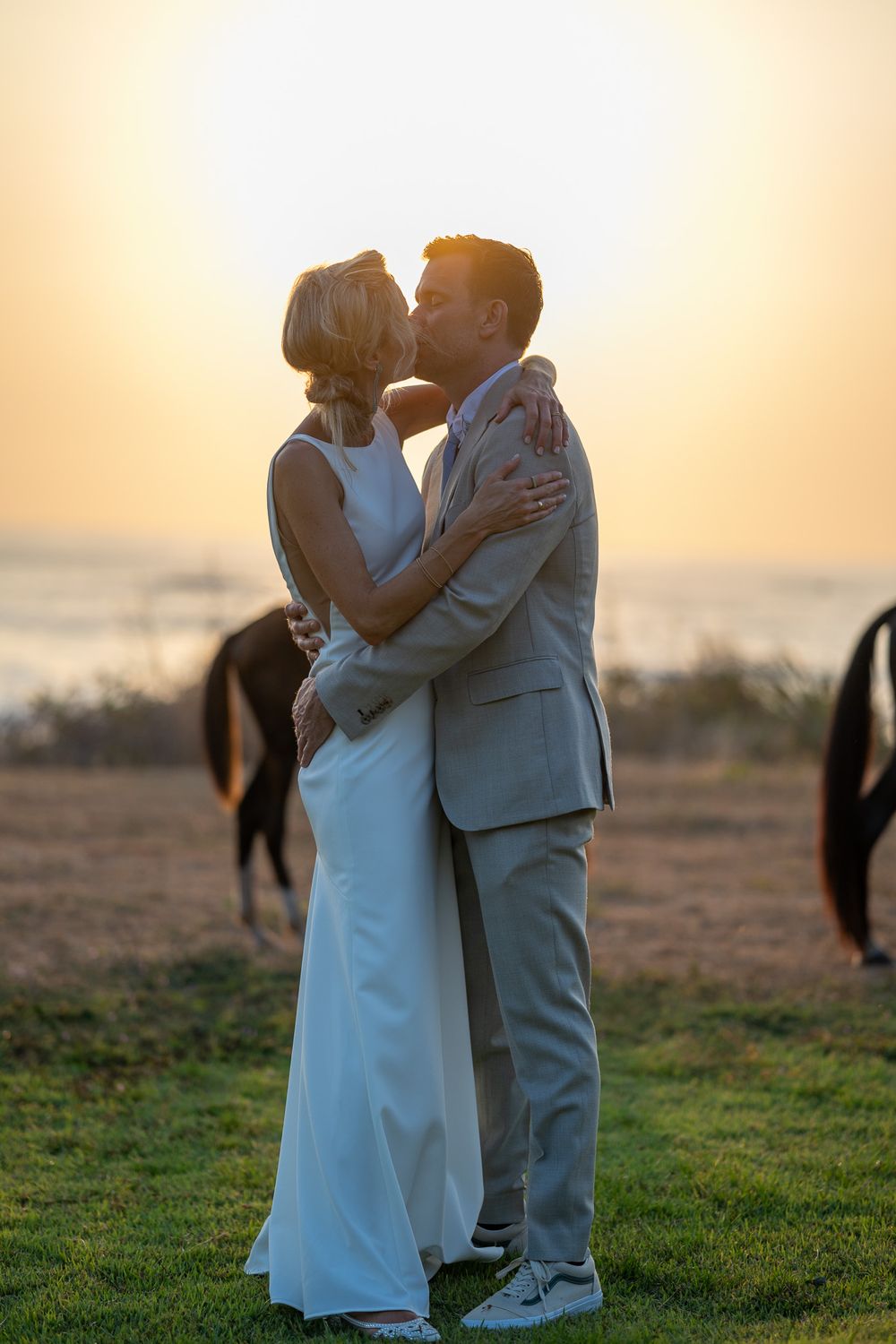Newlyweds share a romantic kiss at sunset with horses in the background.