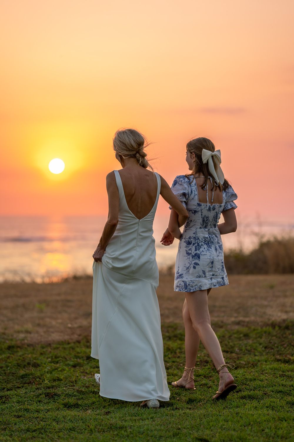 Two women watching sunset on grassy cliff overlooking ocean.
