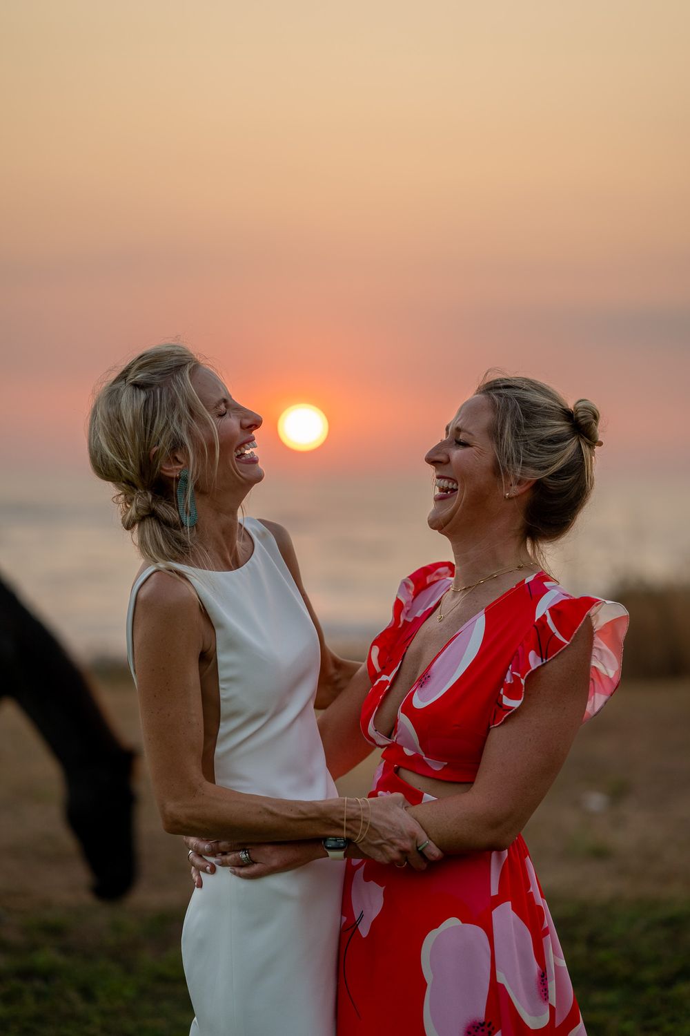Two wedding guests share a joyful moment at sunset by the ocean.