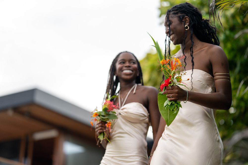 Two women in elegant white dresses hold tropical bouquets while sharing a happy moment.