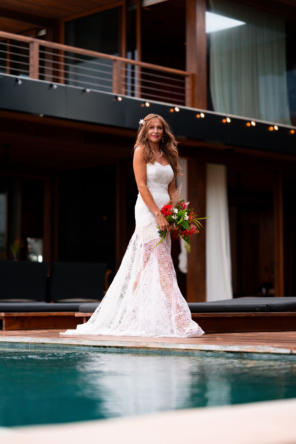 A bride in a lace mermaid wedding dress poses by a modern poolside with tropical flowers.