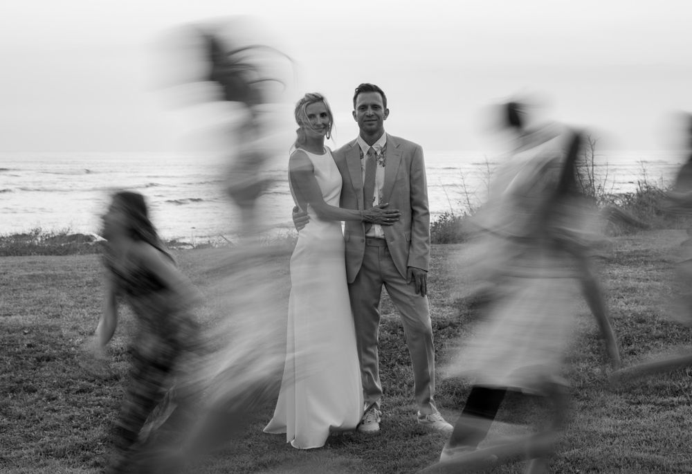A black and white photo with motion blur of wedding guests moving around a still couple.