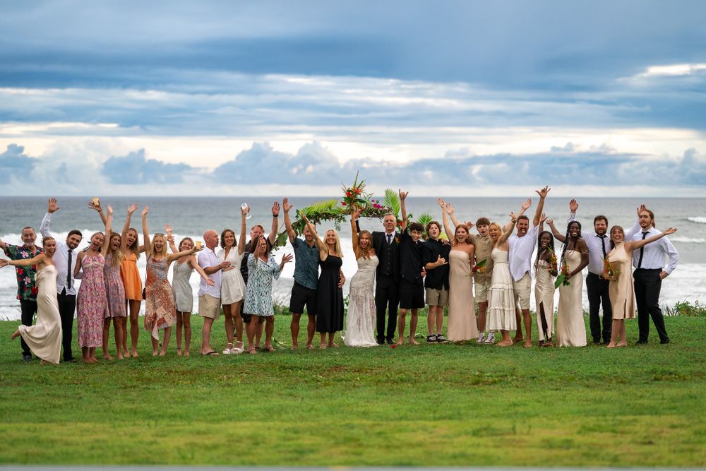 Large wedding party celebrating on a grassy hilltop against cloudy sky with raised arms.