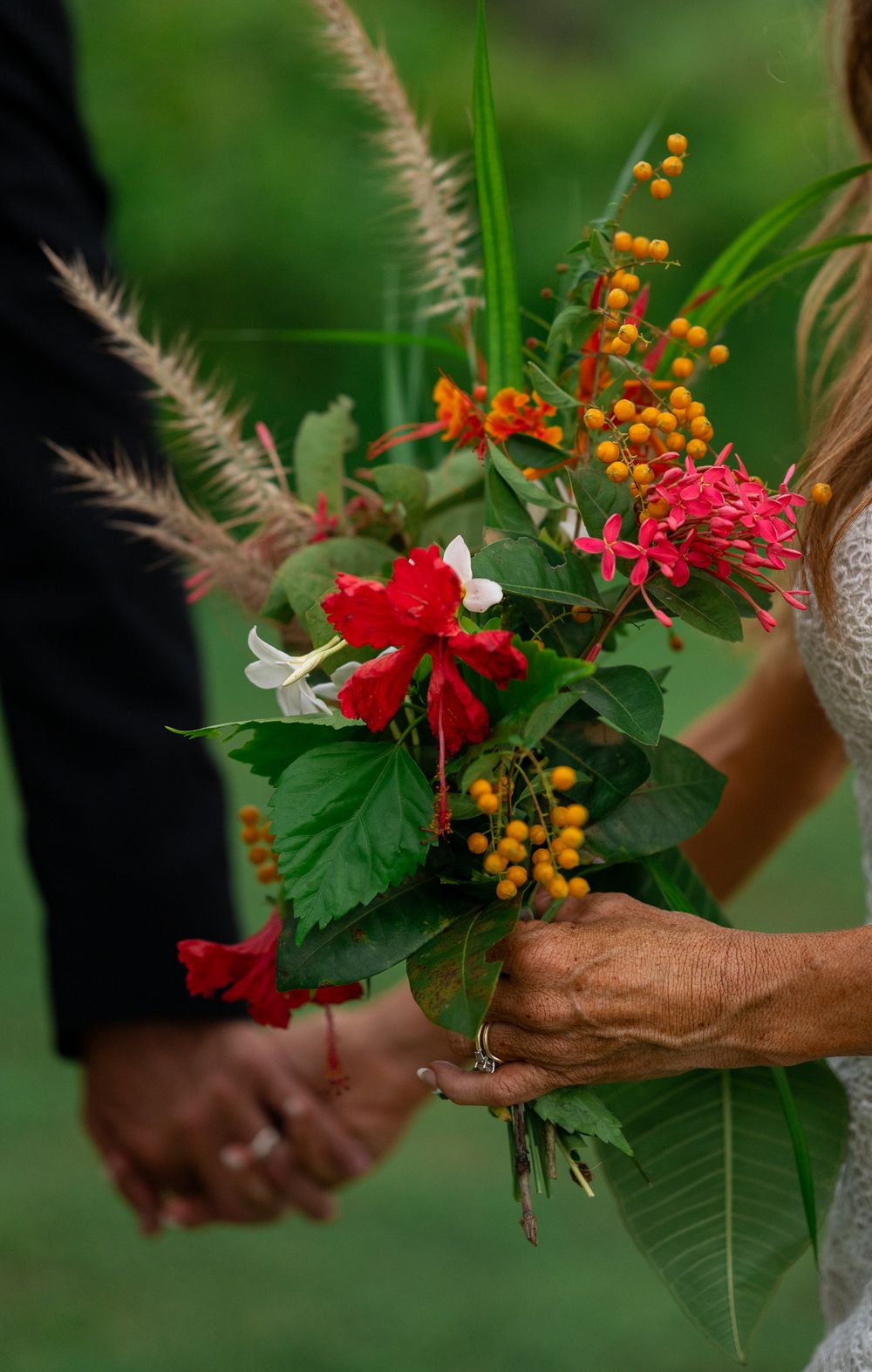 A close-up of vibrant red and yellow flowers in a wedding bouquet held by hands against a blurred green background.