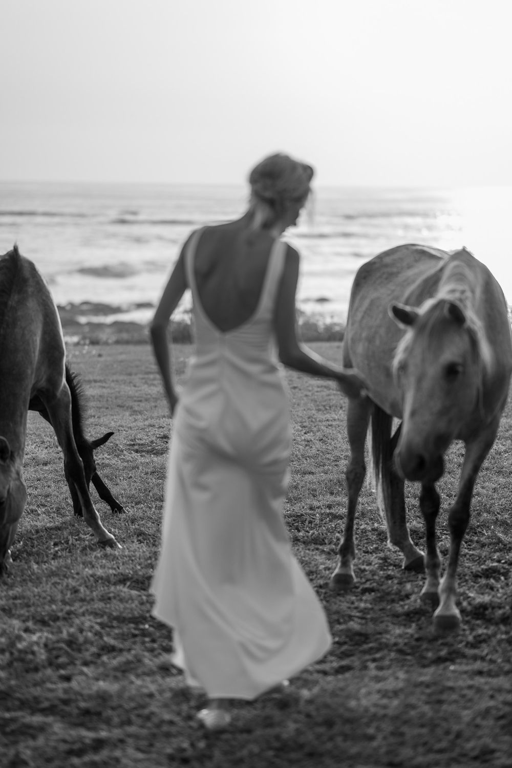 Black and white photo of a bride in a flowing dress walking alongside horses on a beach.