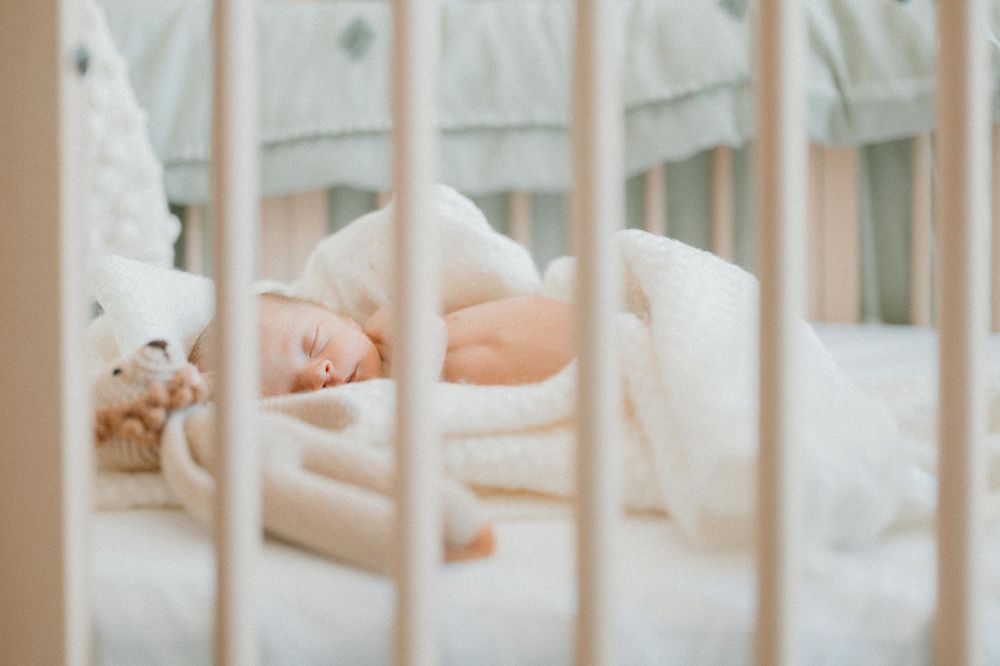 Sleeping newborn baby seen through white crib bars in soft lighting.