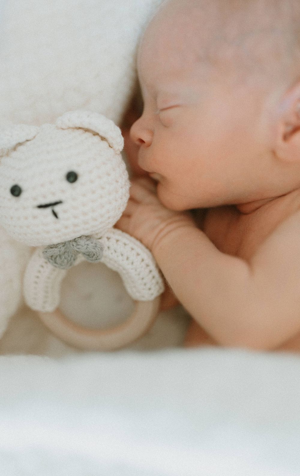 Sleeping baby resting next to white crocheted teddy bear rattle toy.