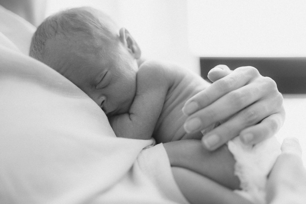 Black and white image of newborn baby resting while adult hand provides comfort.