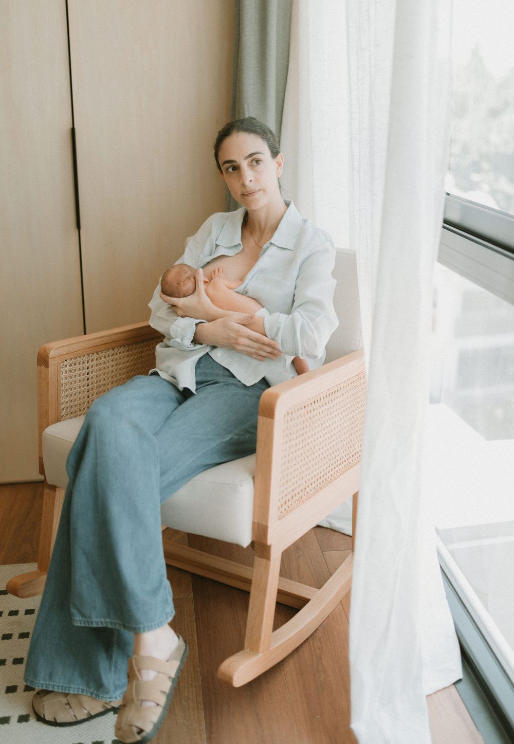 Parent sitting in rattan rocking chair with newborn baby by bright window.