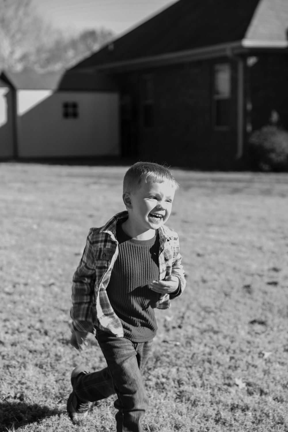 A joyful child in a plaid jacket and overalls runs playfully across a grassy field with a building in the background.