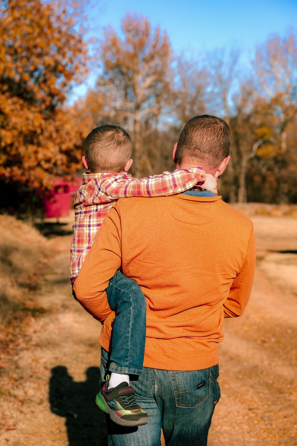 A person in an orange sweater carries a small child along an autumn path surrounded by colorful trees.